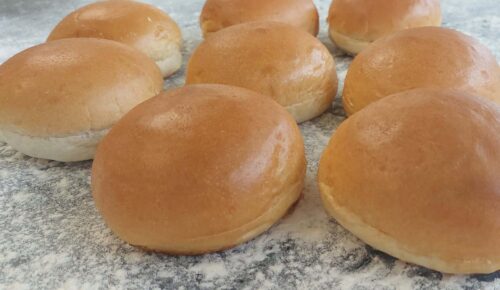 Eight freshly baked brioche buns are arranged on a floured surface. The buns are golden brown and smooth.