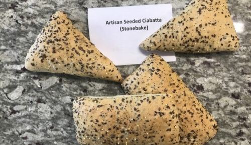 Four pieces of artisan seeded ciabatta, triangular and rectangular, are on a speckled grey countertop with a label reading "Artisan Seeded Ciabatta (Stonebake)."