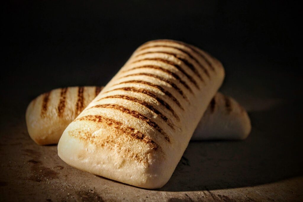 Two golden-brown ciabatta panini breads with distinct grill marks lie on a dark surface.