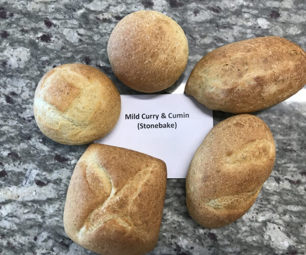 Round and oval bread loaves arranged on a speckled granite surface, with a card reading "Mild Curry & Cumin (Stonebake)" at the centre.