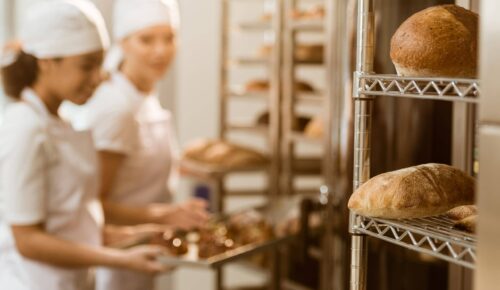 Two bakers in white uniforms and hats stand in a bakery kitchen, placing freshly baked pastries on trays. Shelves hold loaves of bread.