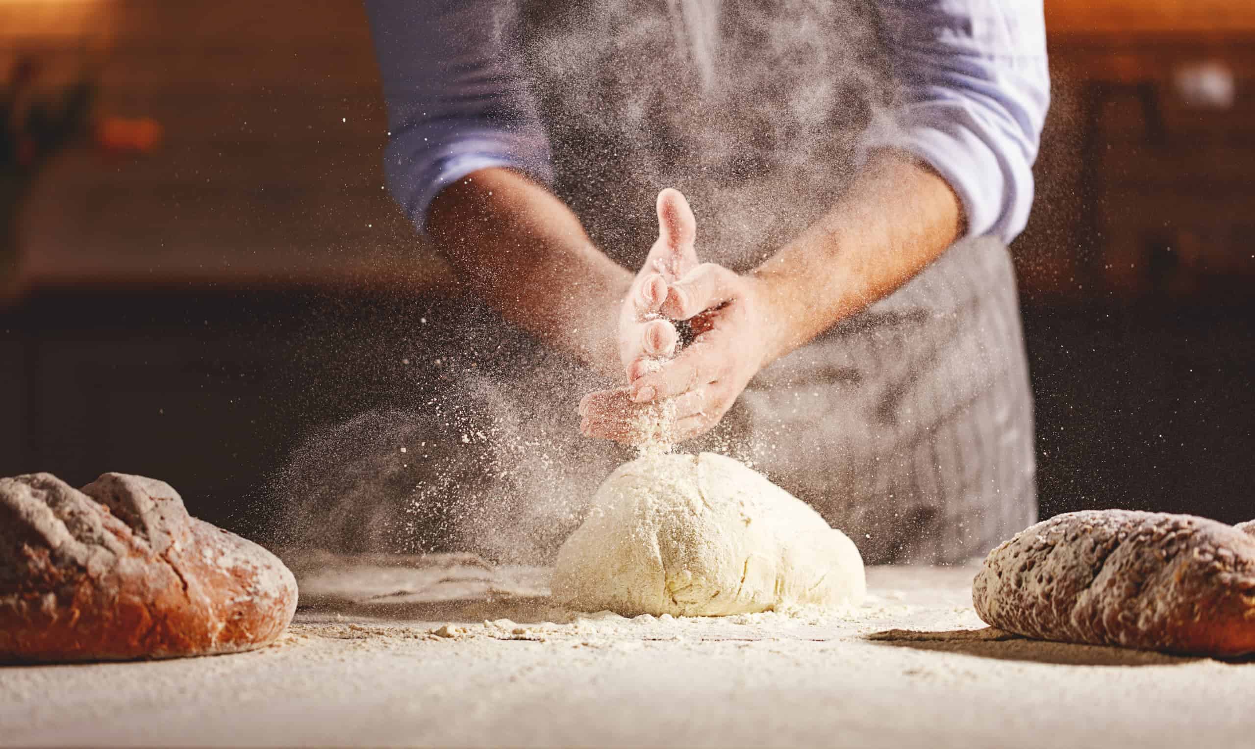 A baker kneads dough, with flour dust in the air, on a wooden table. Two loaves of bread sit on either side.