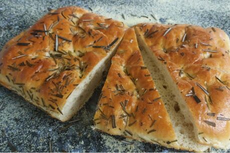 Rustic rosemary focaccia bread, golden brown with visible rosemary sprigs, sliced into pieces on a floured surface.