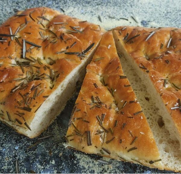 Rustic rosemary focaccia bread, golden brown with visible rosemary sprigs, sliced into pieces on a floured surface.