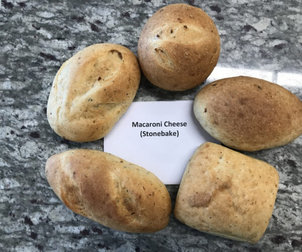 Five round and oval stone-baked rolls are arranged on a speckled countertop. A card in the centre reads "Macaroni Cheese (Stonebake)."