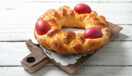 A braided, golden-brown brioche bread wreath sits on a wooden board, adorned with three red Easter eggs.