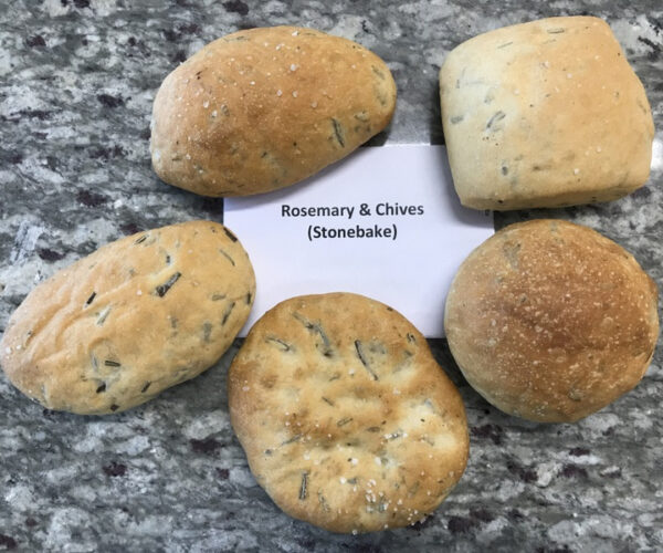 Five freshly baked rolls labelled "Rosemary & Chives (Stonebake)" on a granite countertop. The rolls appear golden and sprinkled with herbs.