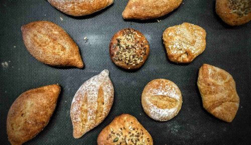 Assorted artisanal bread rolls on a dark baking tray. Breads vary in shape; some topped with seeds.