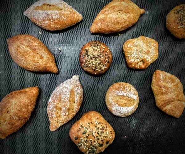 Assorted artisanal bread rolls on a dark baking tray. Breads vary in shape; some topped with seeds.