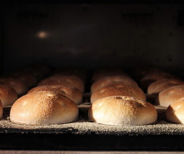 Golden brown buns are lined up on a baking tray inside an oven, bathed in warm light from above.
