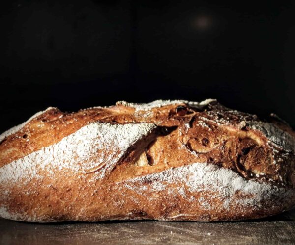 A rustic loaf of bread with a golden-brown crust, dusted with flour, against a dark background.