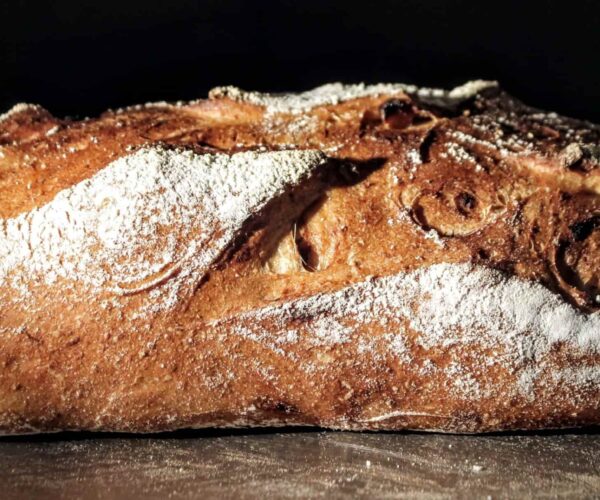 A rustic loaf of bread with a golden-brown crust, dusted with flour, against a dark background.
