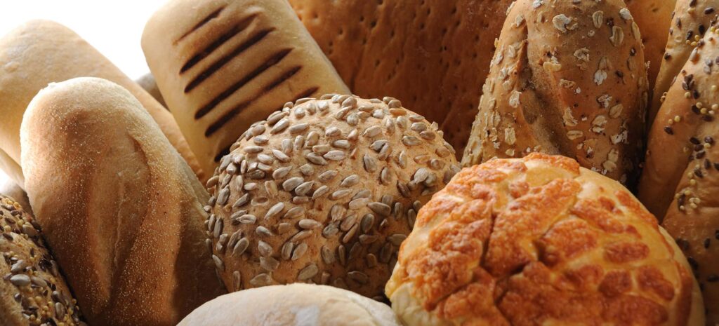 A variety of freshly baked bread, including seeded rolls, crusty loaves, and grain-topped buns, arranged closely to showcase textures and warmth.