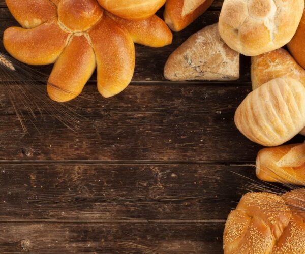 Assorted fresh bread, including rolls and a braided loaf, on a rustic wooden table. Wheat stalks are placed decoratively.