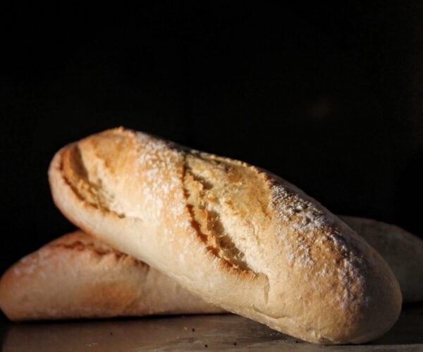 Two rustic, light-brown baguettes dusted with flour are stacked on each other, against a dark background.